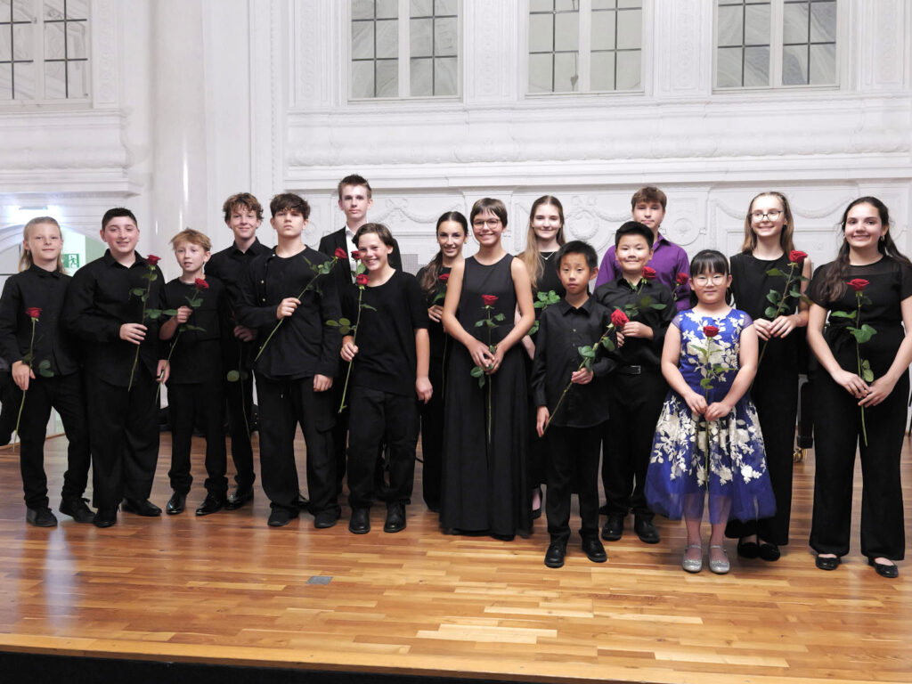 Gruppenfoto der jungen Musikerinnen und Musiker auf der Bühne mit Instrumenten und je einer roten Rose in der Hand